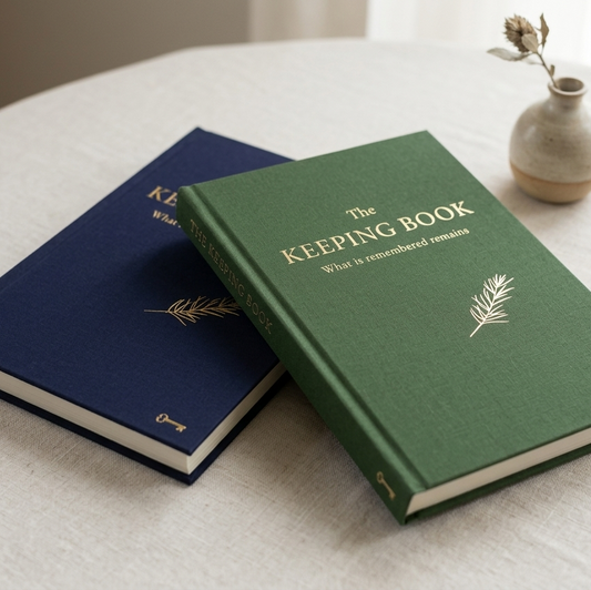 Two hardcover keepsake books, one in dark blue and one in forest green, both with gold lettering that reads 'The KEEPING BOOK' and a small gold botanical illustration. They are styled on a light beige linen-covered round table with a small ceramic vase in the background.